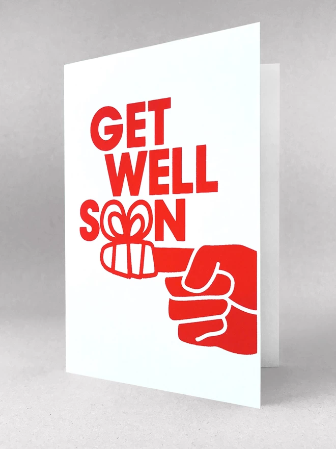 A finger wrapped in a bandage form the two o’s of the word SOON as part of this Get well soon card. Printed in red ink on a white portrait format card. Stood slightly open, in a light grey studio set.
