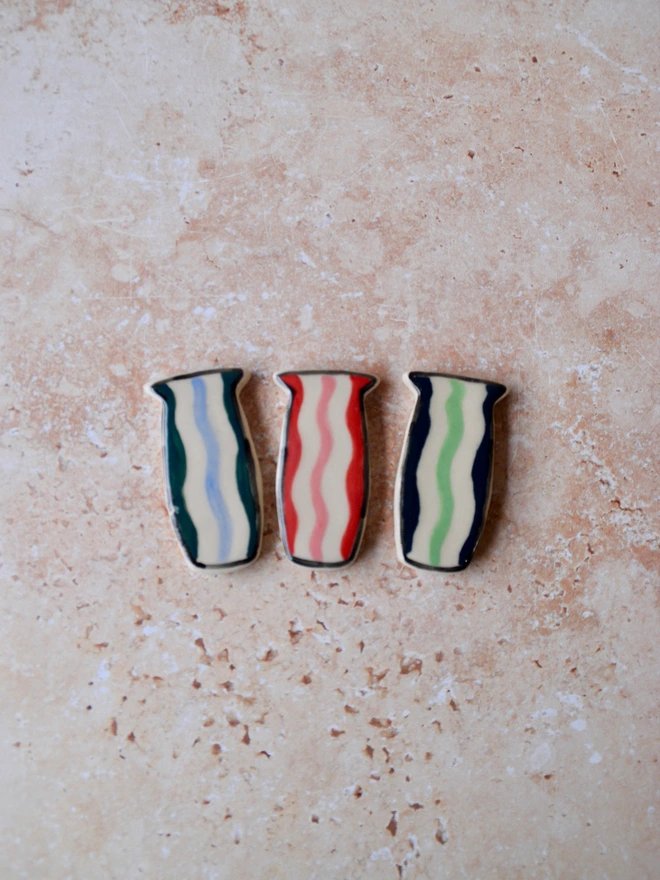 Handmade ceramic magnets shaped like small vases, decorated with wavy stripe patterns in red, blue and green tones, displayed on a textured neutral surface.
