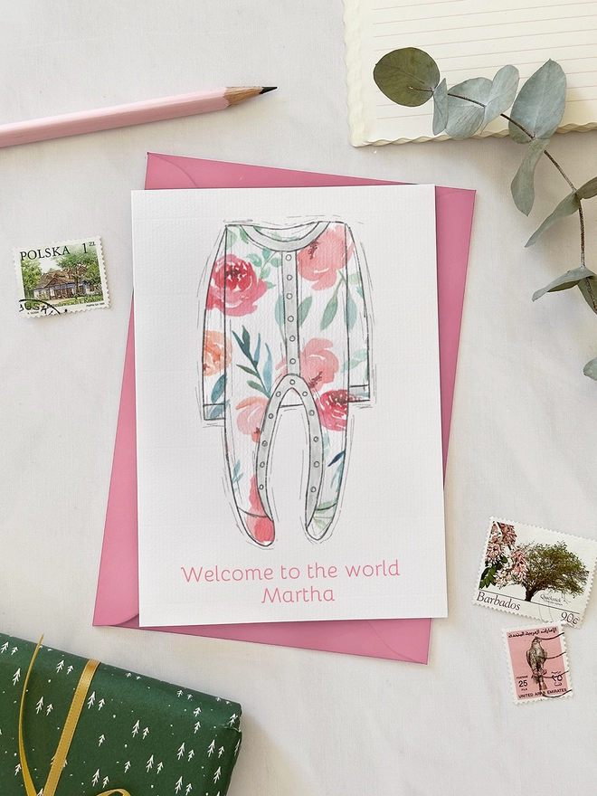 An illustrated new baby greetings card with a pink onesie design lays on a pink envelope on a white desk.