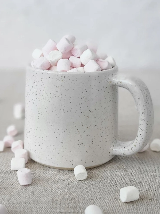Ceramic Standard Mug, a colourful ceramic mug sitting on a plain surface against a grey backdrop. It is accompanied by marshmallows. 
