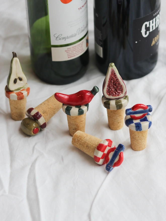 Fig, Olive, Chilli Or Pickle Bottle Stoppers, a group of bottle stoppers on a white table cloth next to some wines. 