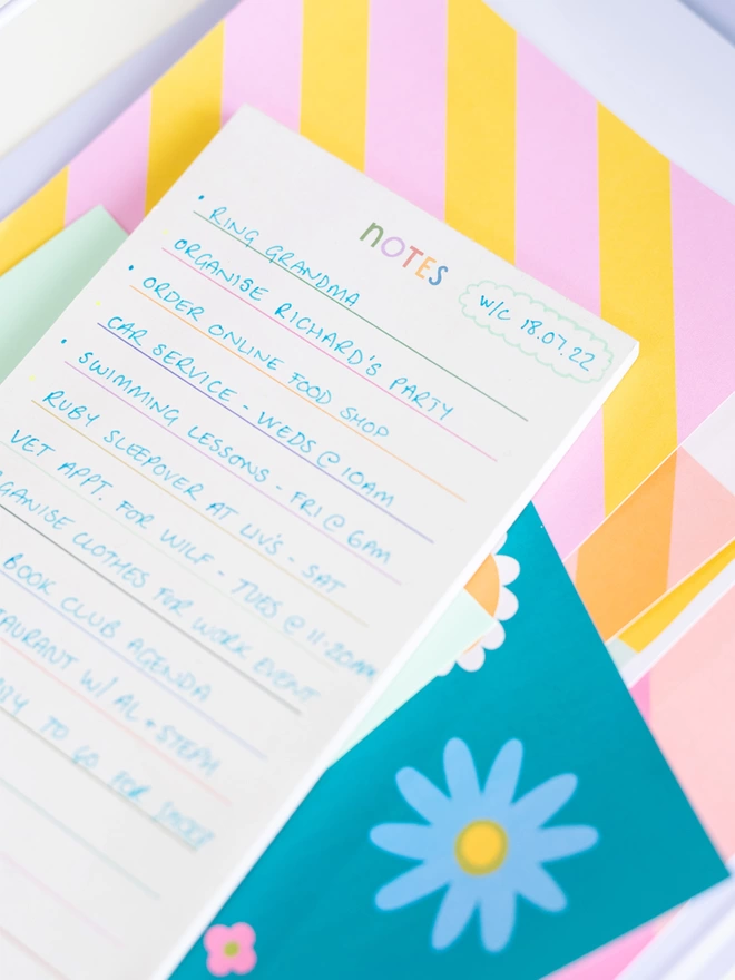 The rainbow lined note pad with a handlettering multi coloured 'notes' title sits on desk with other items from the Raspberry Blossom Happiness stationery collection