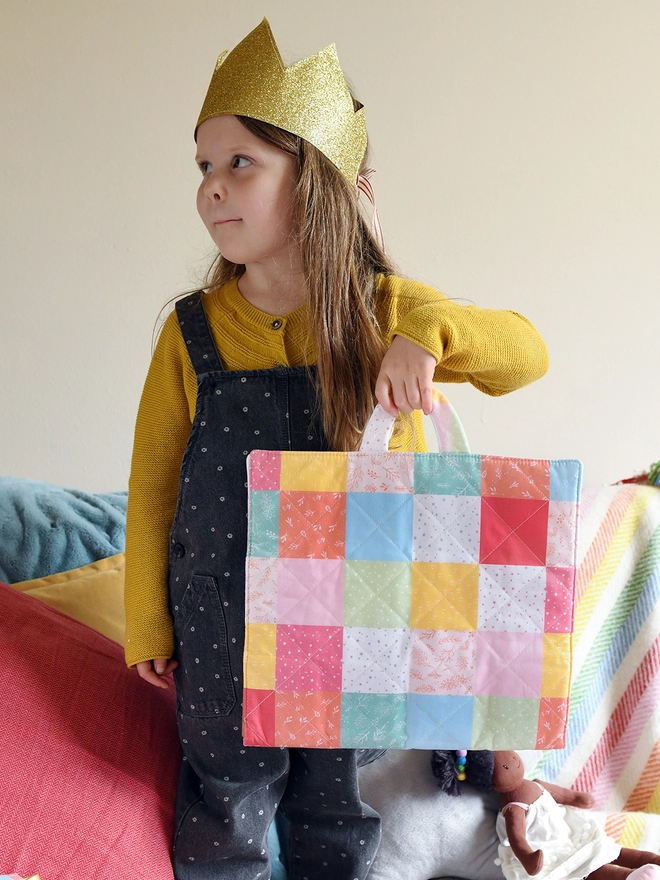 A young child wearing a yellow top and grey dungarees, and a gold crown, stands holding a patchwork quilted folder.