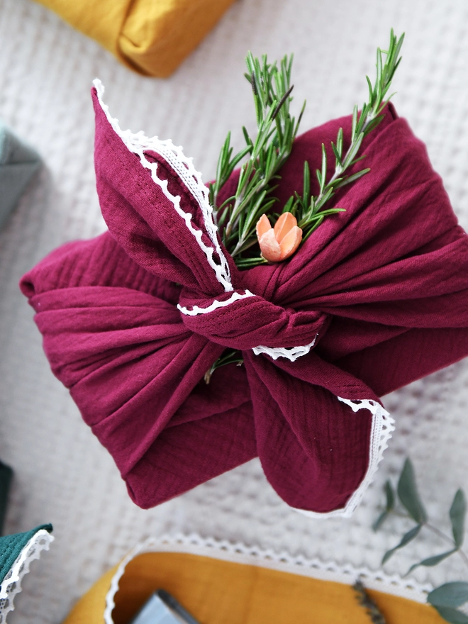 A gift wrapped in burgundy fabric wrap with an ivory lace trim stands among a pile of other wrapped gifts on a white table.