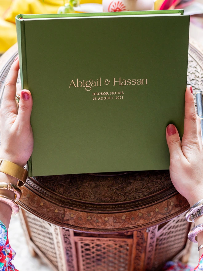 A woman is holding a lovely green guestbook in her hands. The front of the guest book has been personalised with a couple's wedding details.