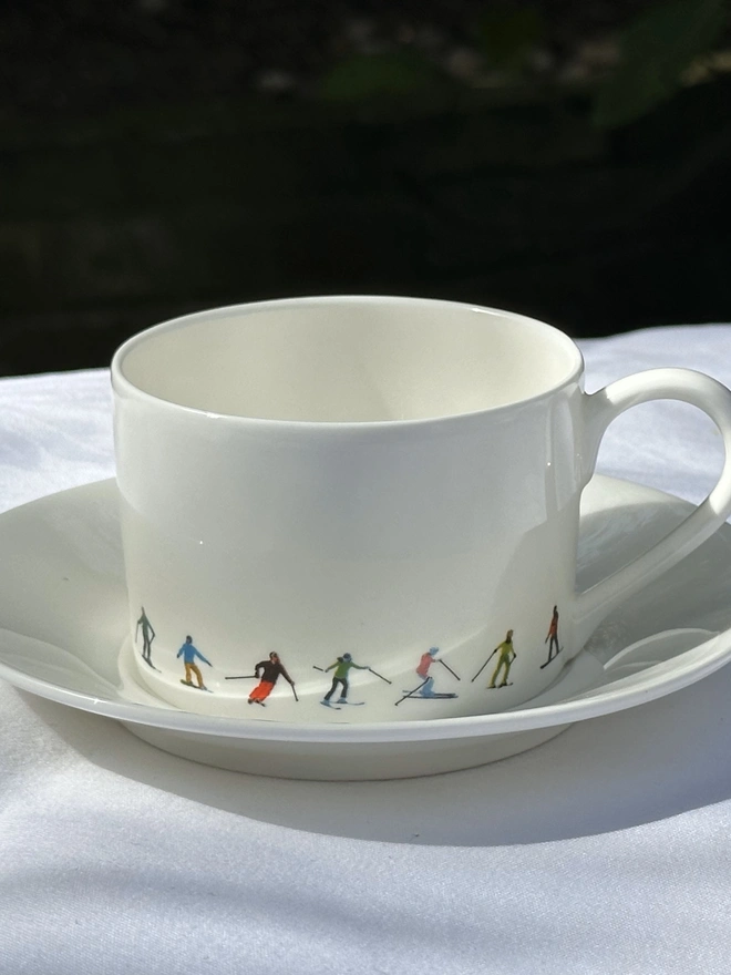 white teacup with chain of multicoloured skiers, and white saucer sitting on white cloth