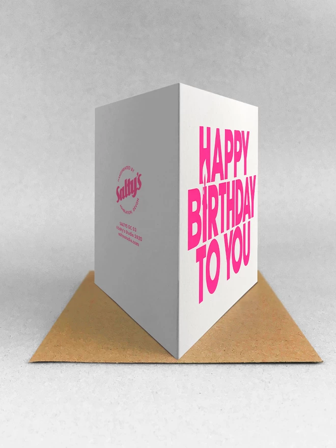 rear view of the neon pink happy birthday screen printed card, stood on a Kraft brown envelope in a light studio shot. 