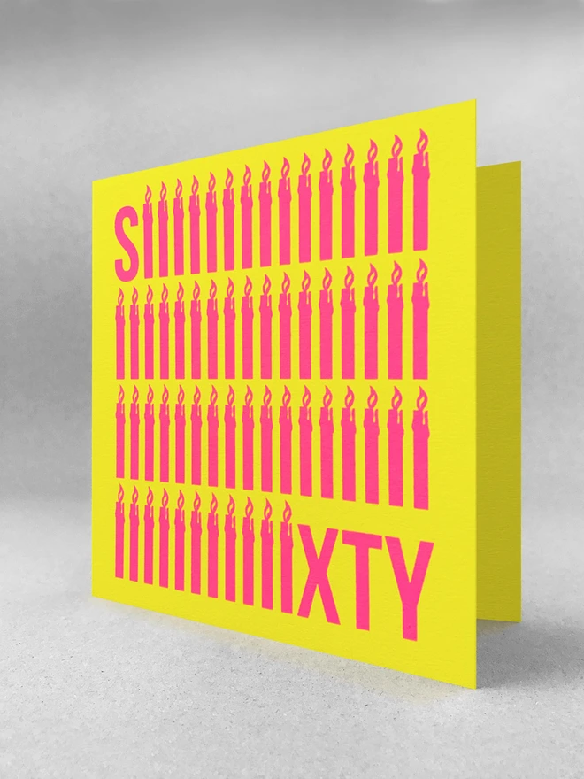 The word Sixty screen printed in neon pink ink across a bright yellow square card.  To emphasise the sixty, the letter i is shaped like a simple  lit candle and is repeated sixty times in four rows. The card is stood slightly open, in a light grey studio set.