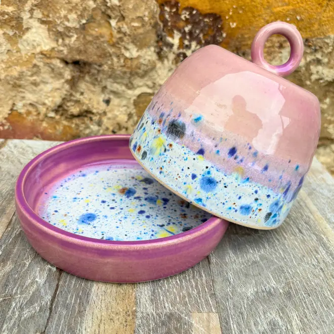 Shoreline Butter Dish, a colourful butter dish placed on a wooden surface against a brick wall. The lid is coming off in the image. 