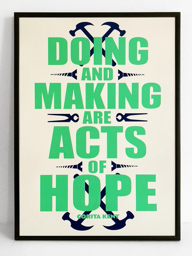 Framed green and blue typographic print. The Corita Kent quote reads "Doing and making are acts f hope." Navy blue tools are placed around the green text.