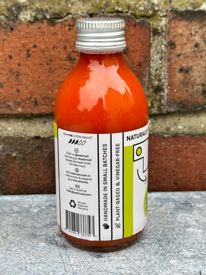 Picture Of A Muti Troublemaker Bottle Facing Left And On A Steel Topped Table Against A Red Brick Wall.