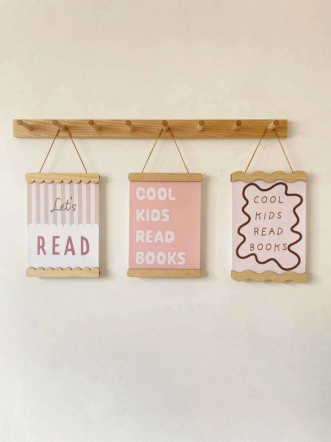 Three wooden picture frames hung on a peg rail. the picture frames are straight, wavy and scalloped in style. Each is strung with vegan suede and holds children's colourful prints reading Cool Kids Read Books and Lets Read