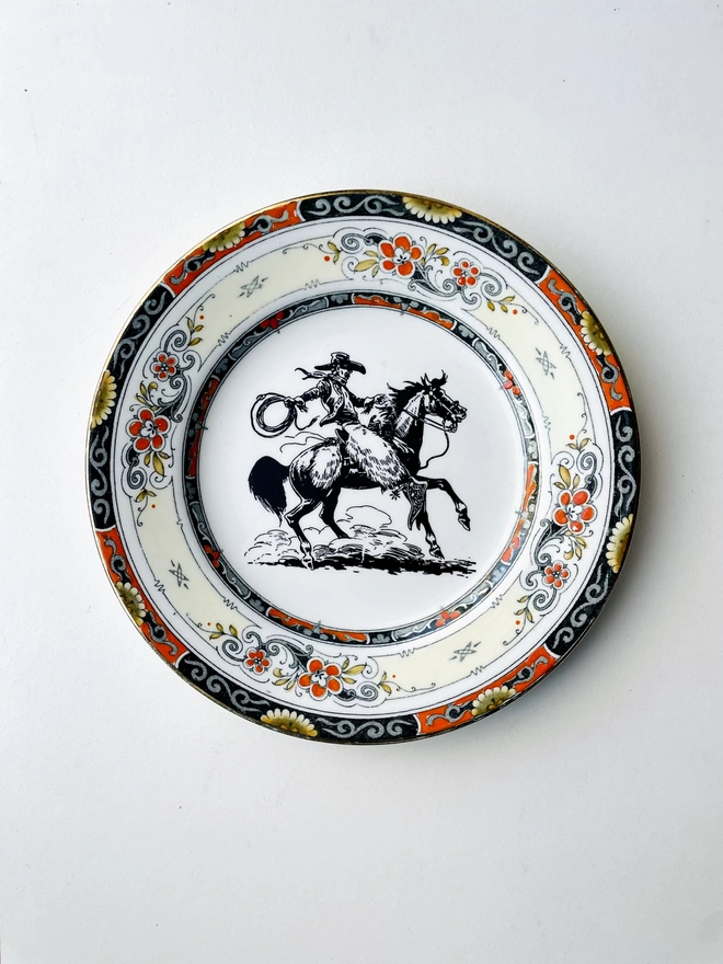 really decorative vintage plate measuring 17cm across, it has a really ornate border in orange black white and cream pattern, and in teh centre of the plate is a vintage black white illustration of a cowboy on his horse