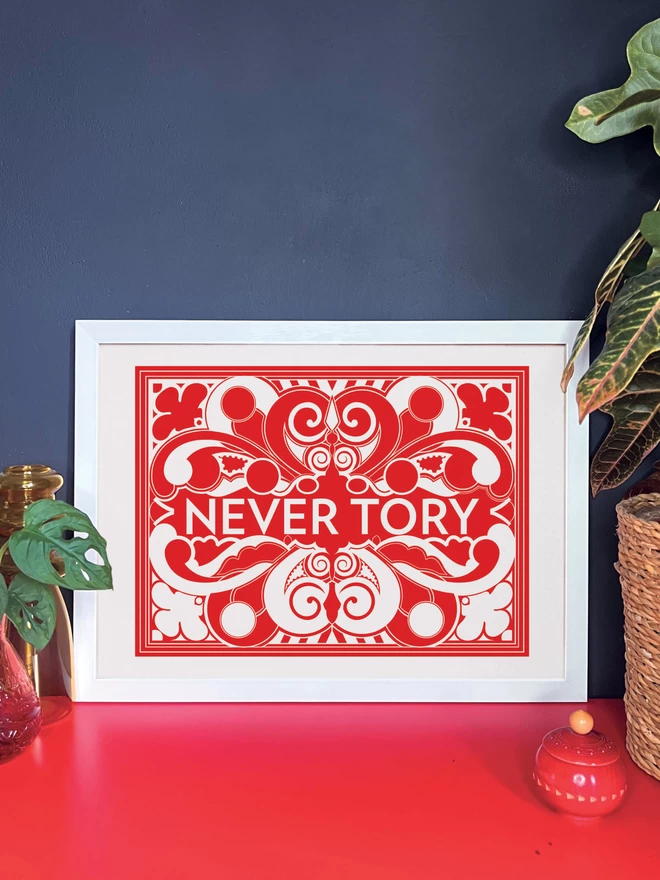 A bold, landscape red and white symmetrical illustration with Never Tory written in white at the centre. The print is in a white frame resting against a dark grey wall on a red cabinet. Next to the frame are two pot plants and a small red wooden pot.