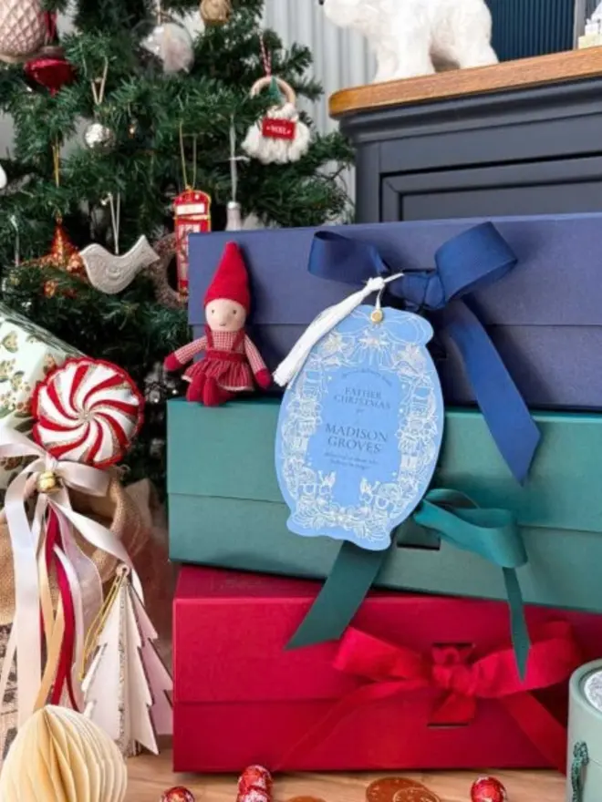 A personalised pale blue gift tag with a silver nutcracker and reindeer border and a white tassel, reading "Father Christmas for Madison Groves". It hangs from a navy ribbon on a stack of navy, green and red gift boxes beside a Christmas tree, an elf doll and a peppermint ornament.