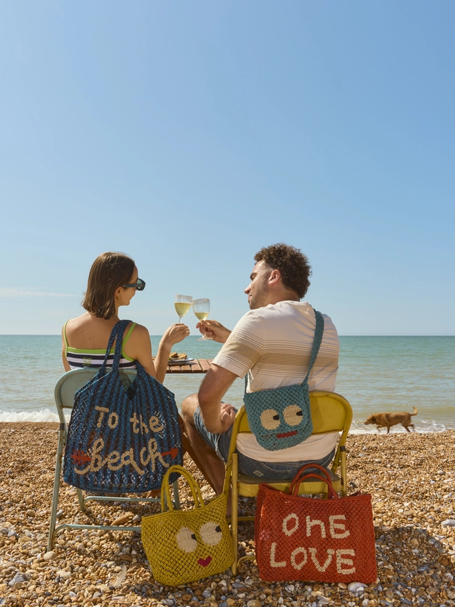 A couple sits on a pebble beach under a clear blue sky, toasting with drinks. Their colorful bags read "To the beach" and "One Love," evoking a joyful, relaxed vibe.