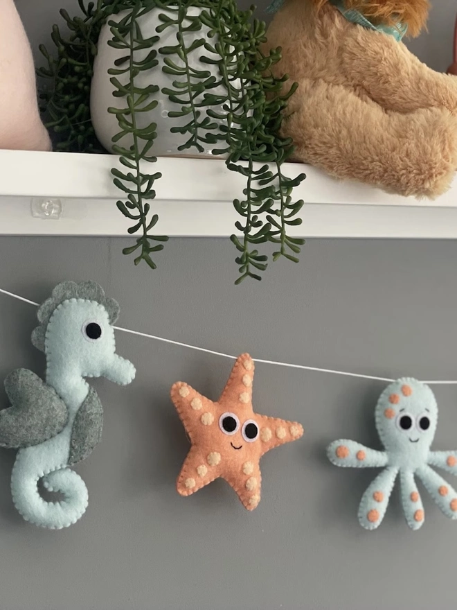Nursery Under The Sea Garland, a sea themed garland hanging from a white shelf against a grey backdrop in a child's bedroom. 