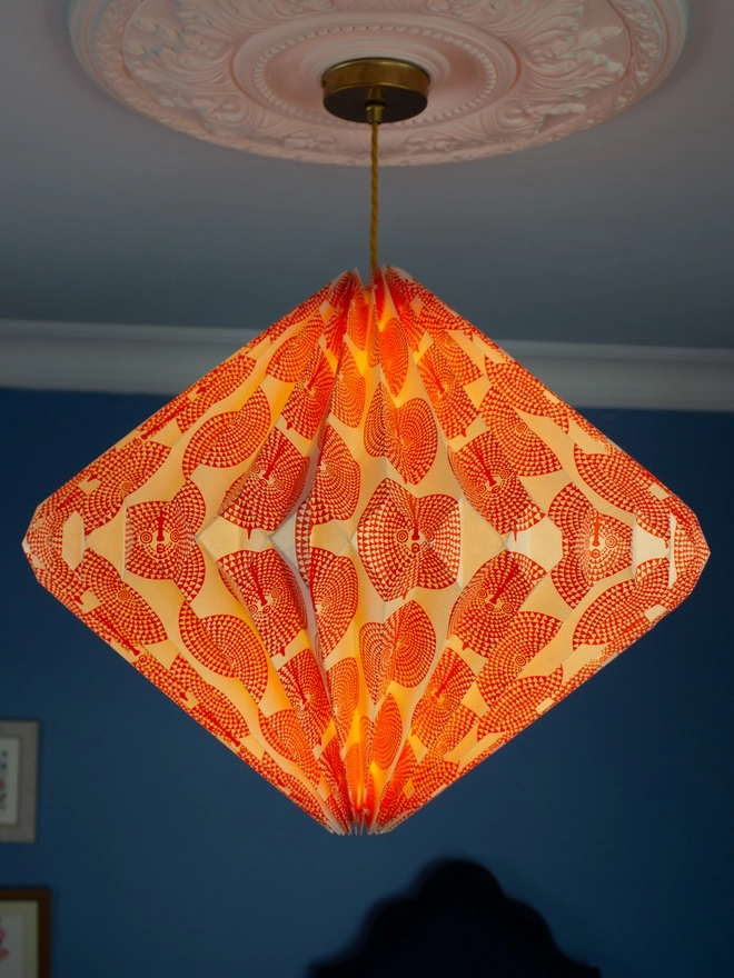 Origami paper lightshade with orange design lit up. Hanging against a blue wall background.