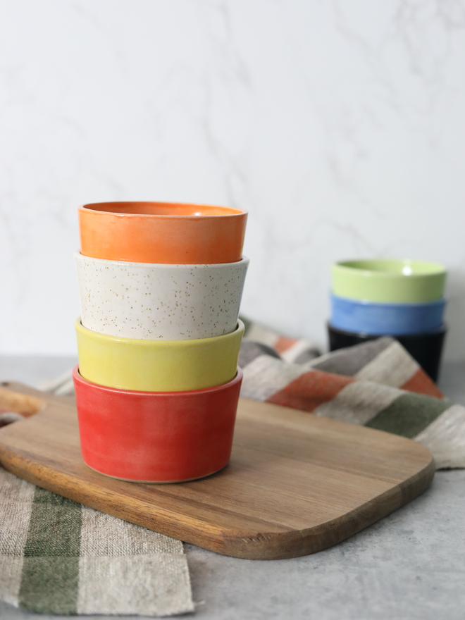 Two stacks of handmade ceramic side bowls on a wooden board — a foreground stack in mandarin orange, speckled white, yellow, and cherry red, and a smaller background stack in spring green, blue, and black — styled with linen cloths against a white marble background