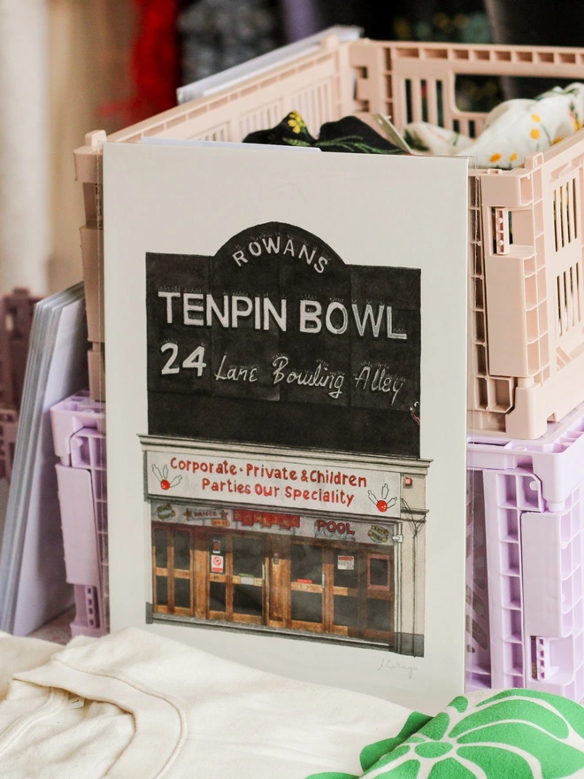 A watercolor print of Rowans Tenpin Bowl leaning against pastel storage crates next to folded fabrics.
