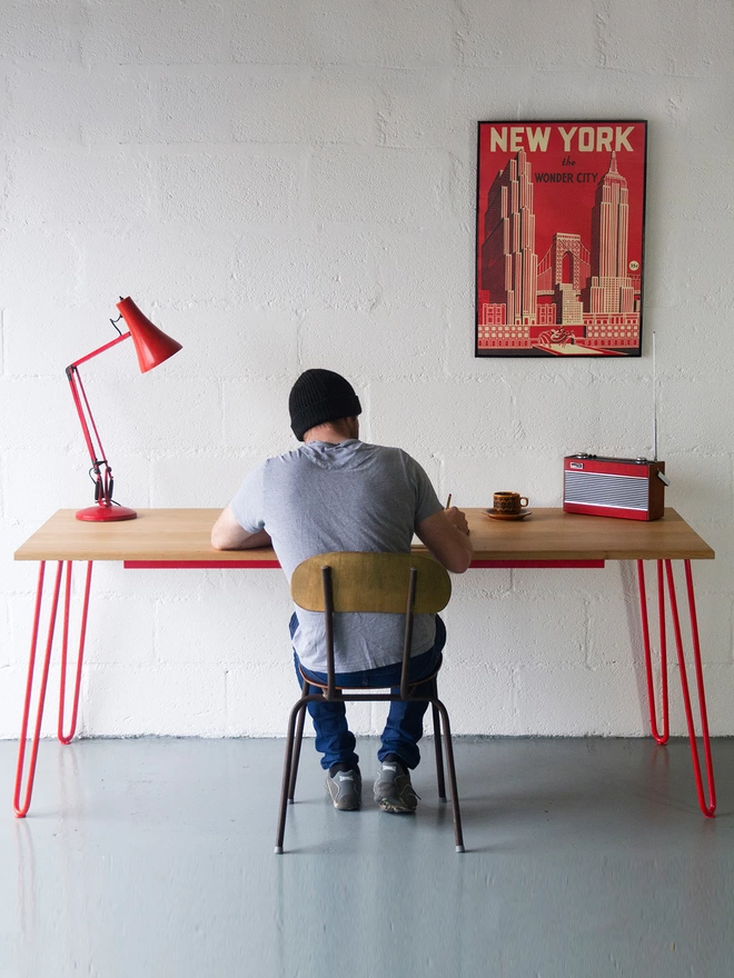 man working at a hairpin leg desk with oak top and red legs, with a red anglepoise lamp and red print on the wall