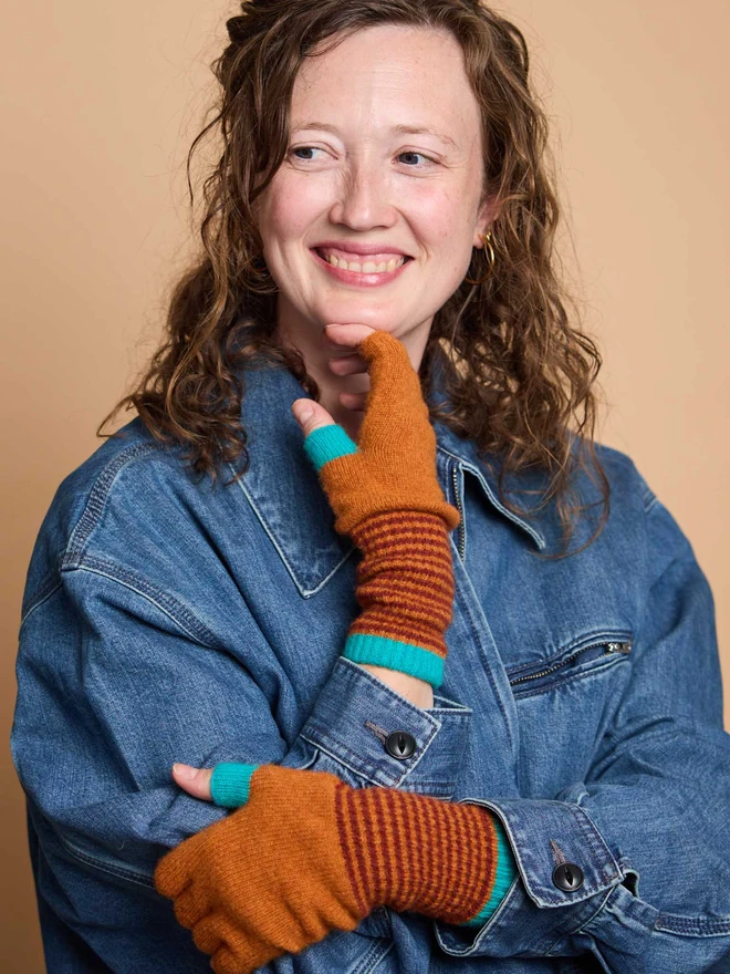 Woman Wearing Rust Striped Fingerless Gloves In A Blue Jacket In front Of A Peach Background