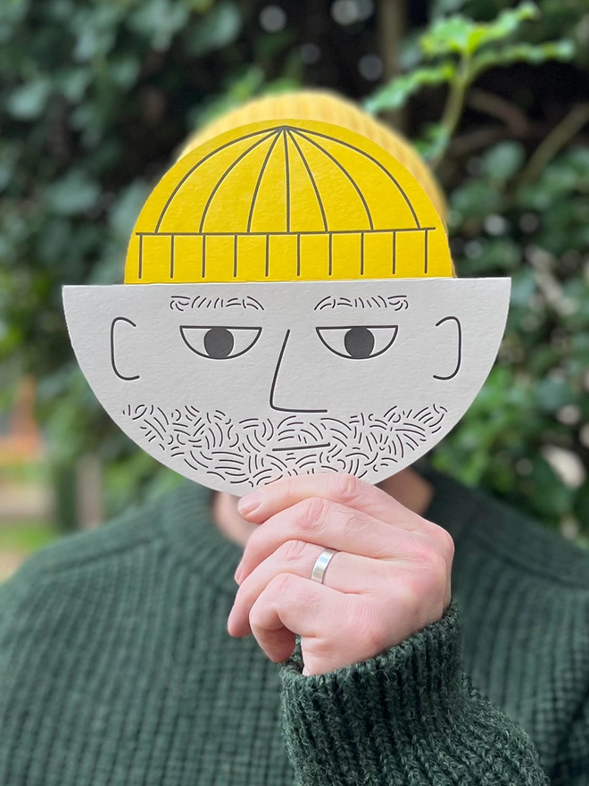 Man with yellow hat holding a round card with a male bearded face and yellow pop up hat. 