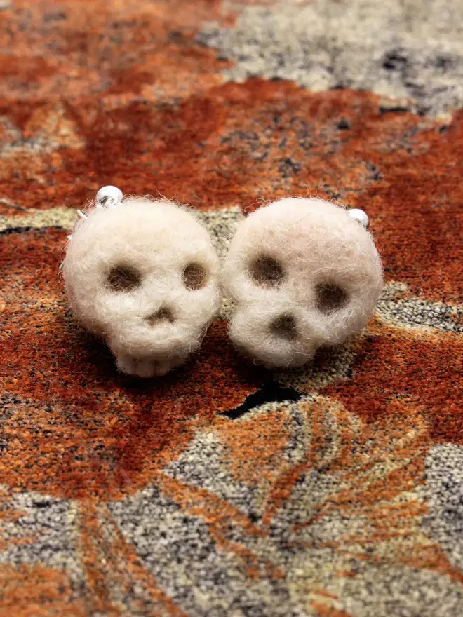 A pair of needle-felted skull earrings on an orange autumnal fabric.  Made by the artist Laura Cronin aka Bumble & Earwig