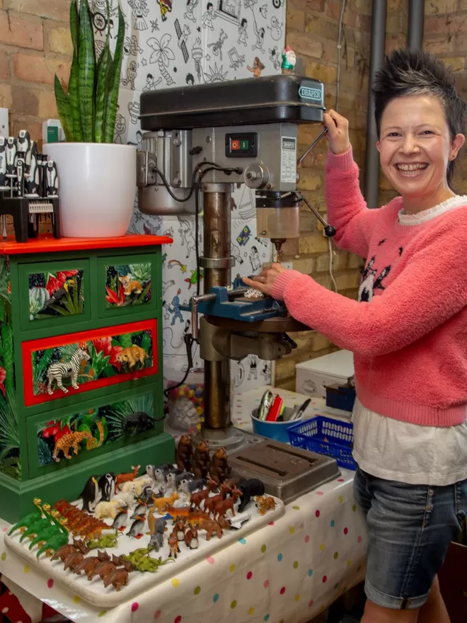  A white female known as the Candy Queen stands to the right of a pillar drill on a garage workbench, she faces the camera smiling, to the left of the pillar drill there is a small chest of drawers with animal knobs attached, there is a tray of plastic toy animals on the workbench.