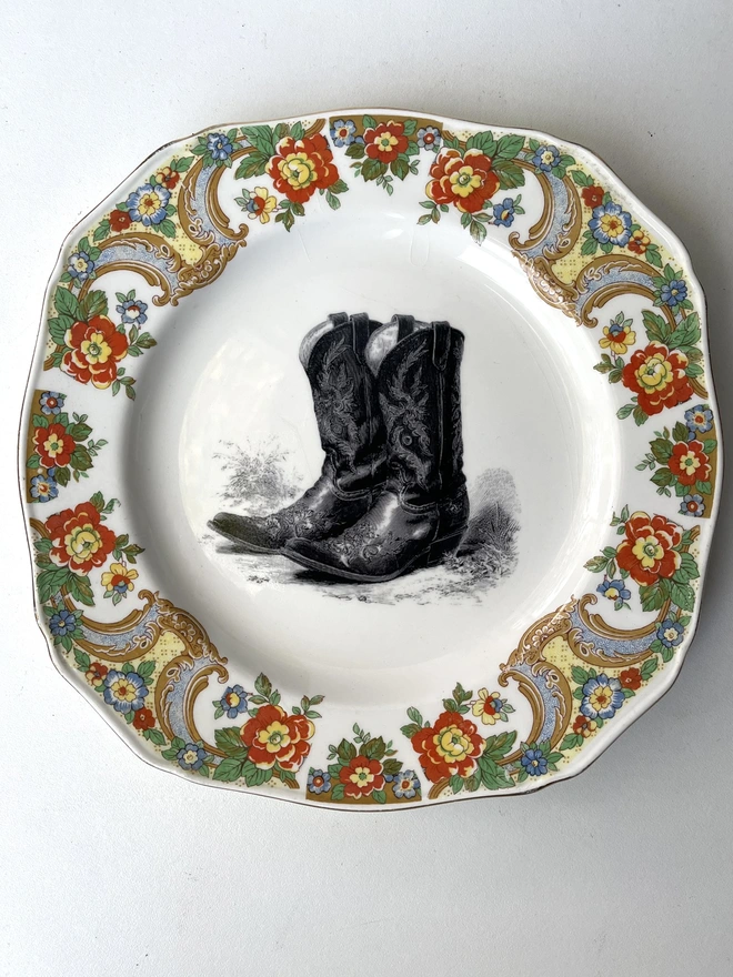 a vintage plate with an ornate border has a black and white vintage illustration of a pair of ornate cowboy boots in the centre