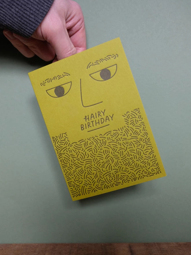 Man holding a 'kiwi' coloured card with a simple beaded face and the text 'Hairy Birthday'.