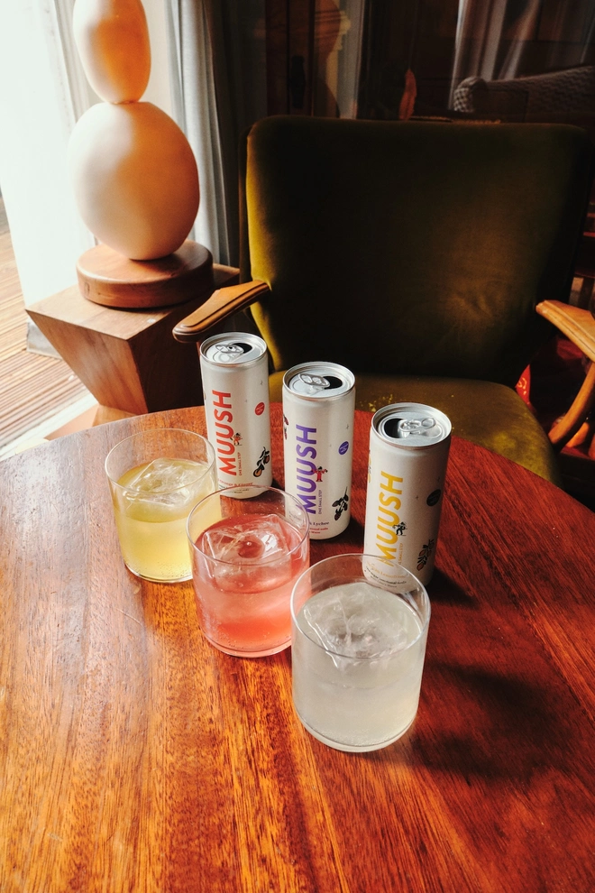 three cans of MUUSH lion's mane soda next to glasses filled with each flavor, on a wooden table