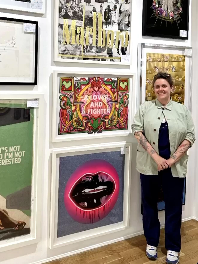 Rebecca Strickson stands next to a framed “A lover and a fighter” illustration hung on a busy art gallery wall. 