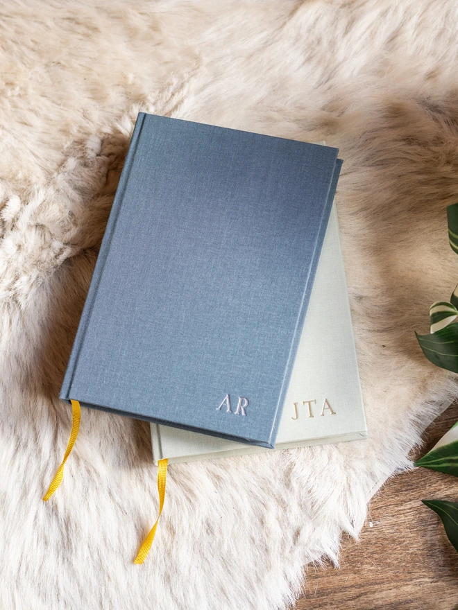 A pile of linen-bound diaries, personalised with printed initials, with a pen resting on top.
