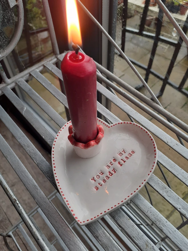 Close-up of a handmade heart-shaped ceramic candle holder in glossy white with red dotted edging and the phrase ‘You are my steady flame’ stamped in red.