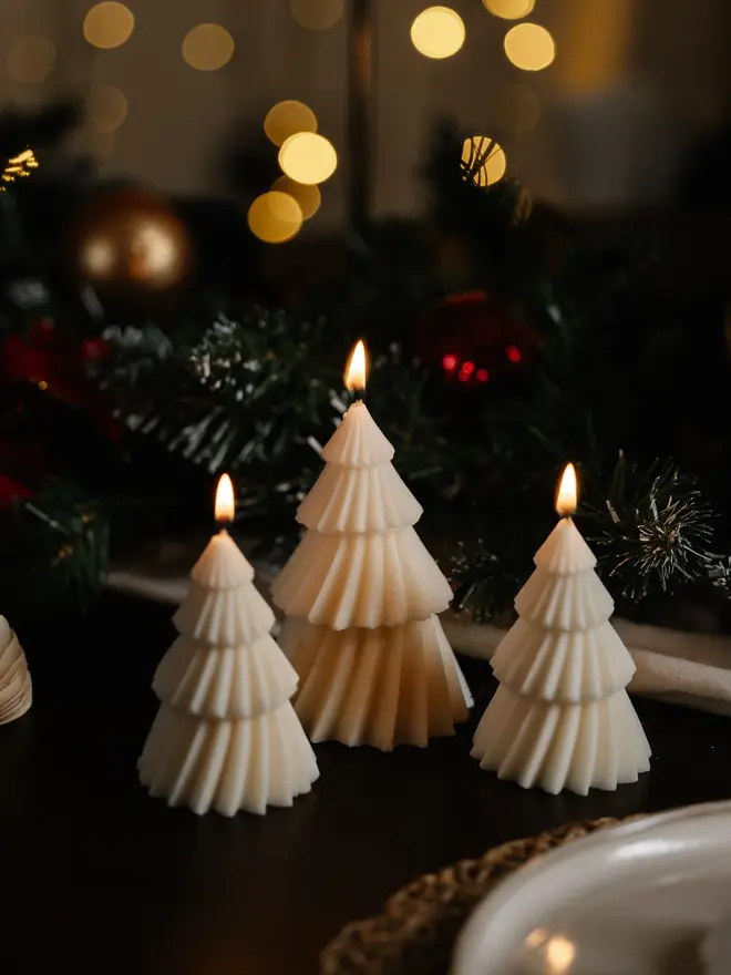 White Christmas Tree Warm Candles (Set Of 2). Three lit cream Christmas tree candles glowing softly beside frosted pine garland and festive baubles. The warm candlelight shows off the crisp ribbed tiers, ideal for creating a magical Christmas atmosphere.
