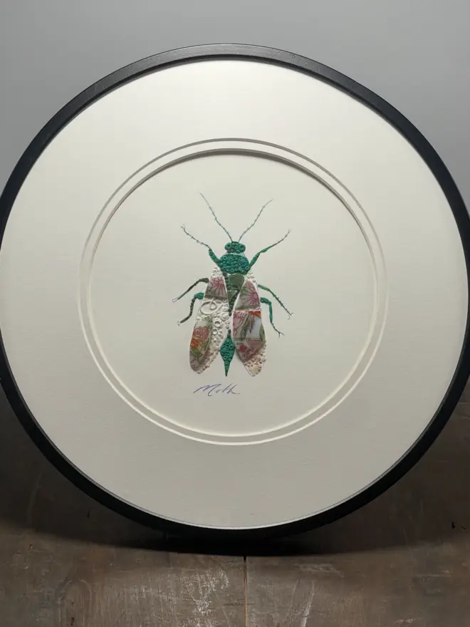 Hand-drawn, painted, mosaic clay, vintage pink floral pottery insect on white double mounted card, in a black circular frame.