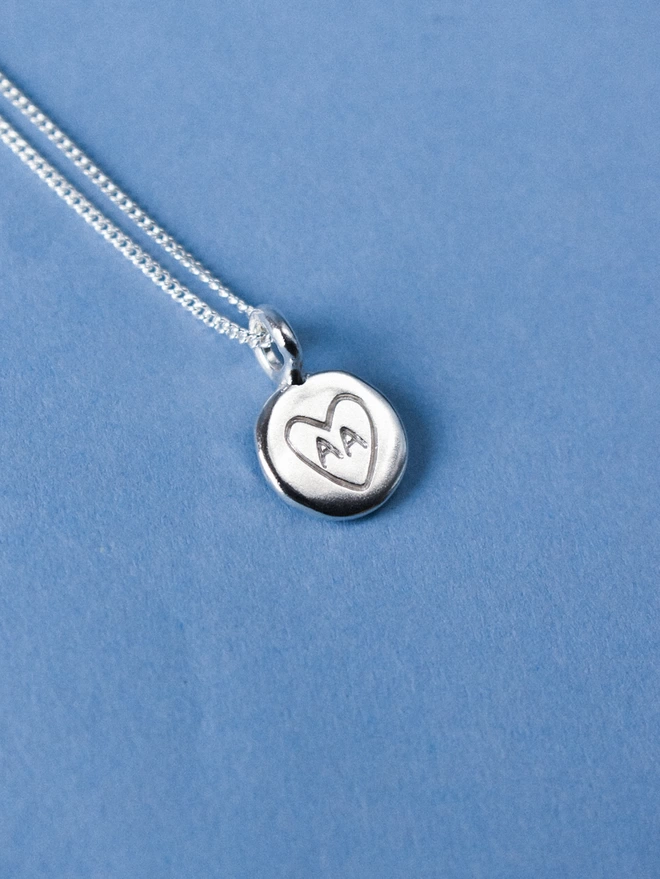 a silver disk pendant with a hand engraved heart with 'AA' in the centre, the pendant is on a. thin silver chain and lays on a pale blue background