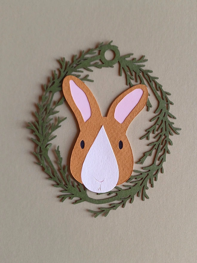 Black Bunny Easter Tree Decoration. Toffee brown bunny face with white nose patch, black eyes and pale pink ears inside olive green foliage wreath oval