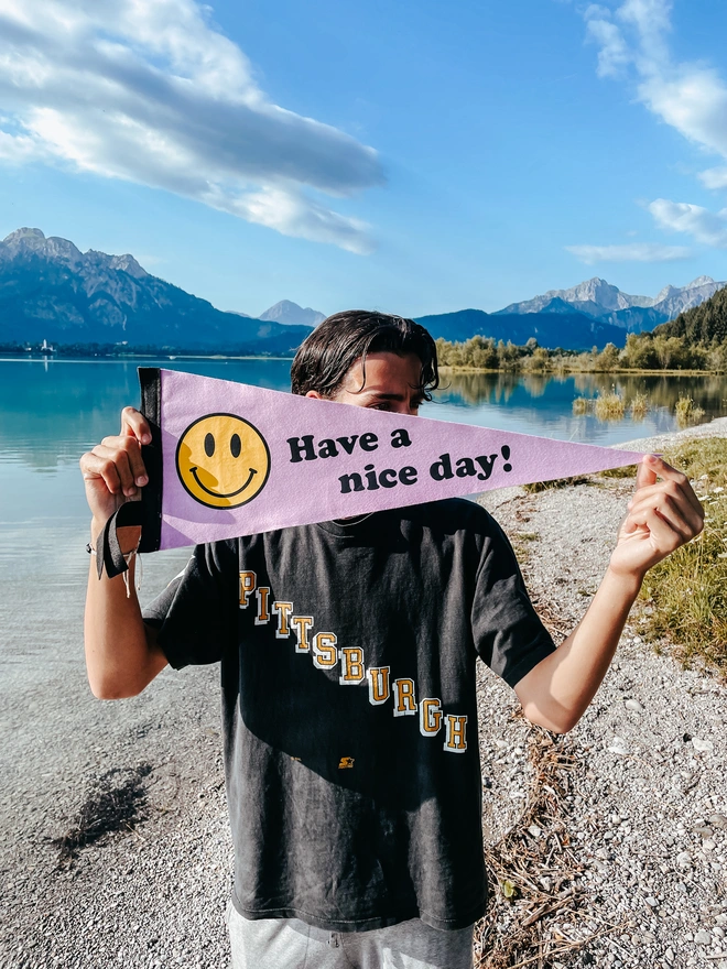 a man stands by the edge of a turquoise lake he holds up a pennant flag that is lilac with a yellow smiley face on, adn teh words have a nice day
