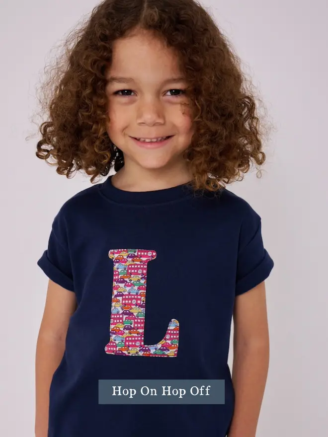 a 5 year old boy wearing a personalised navy t-shirt with his initial cut from liberty london buses and cars print on the front