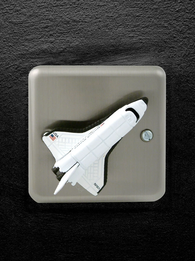 A brushed chrome dimmer light switch with a space rocket as the rotary knob to turn the lights on and off on a black background. The children's light switch brand is Candy Queen Designs.
