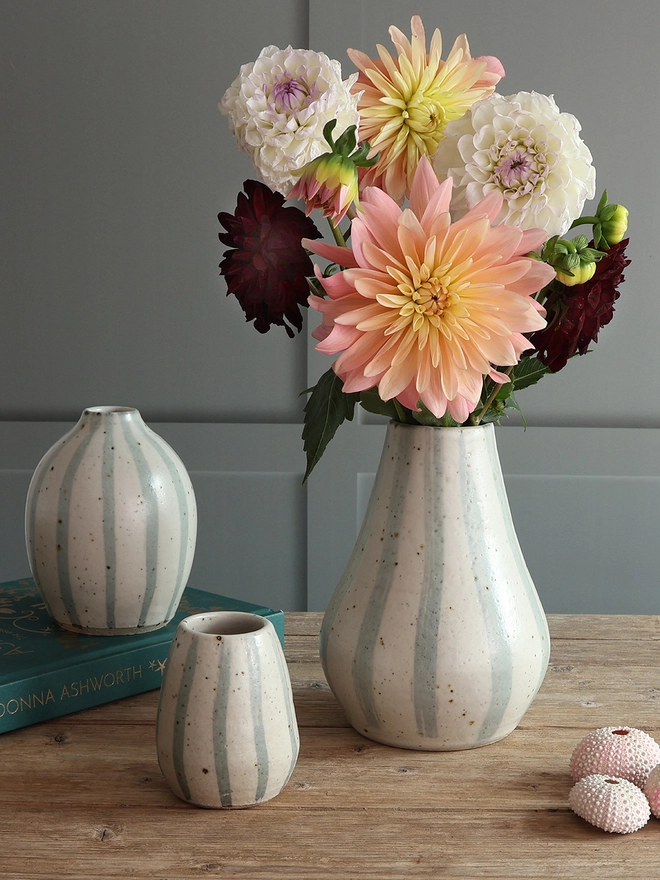 Handmade Ceramic Stoneware Vases in three sizes: small, medium and large. The vases sit on a wooden surface alongside a book and pink sea urchins. The largest vase is filled with a large bouquet of flowers. 