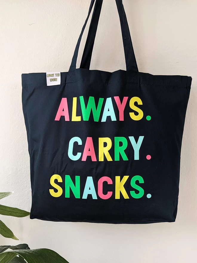 A hanging navy canvas tote bag with Always Carry Snacks slogan