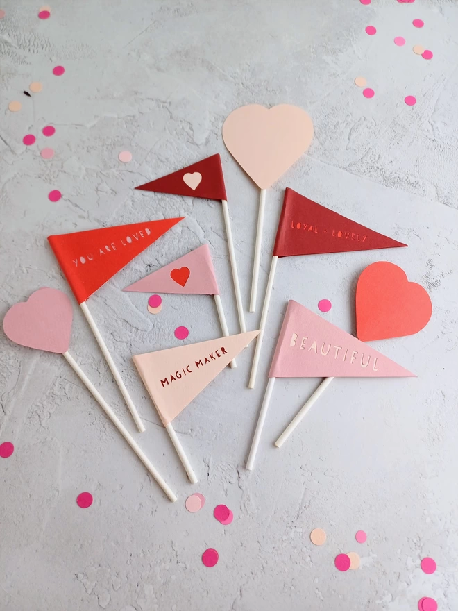 9 paper decorations in shades or red and pink,  attached to paper lollipop sticks. 3 different sized hearts, 2 triangular heart shaped flags with little hearts in the centre, 4 triangular flags each carrying a different word or phrase.