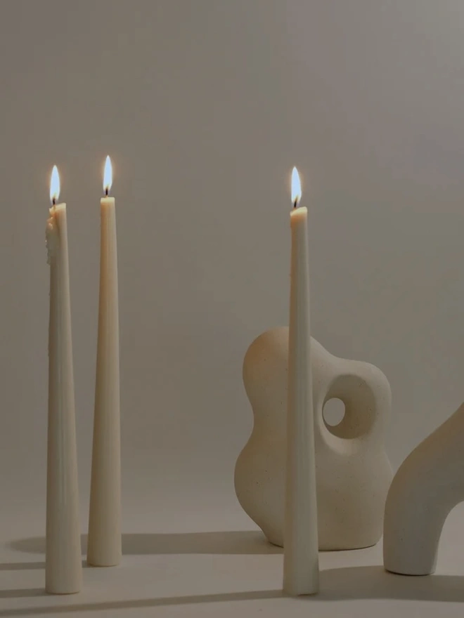 White Stem Candles (Set Of 3). Three lit cream taper candles with tapered bases, standing beside sculptural speckled stoneware ornaments against a soft neutral wall. The elegant, self-standing candles bring a calm, warm glow to modern living spaces.