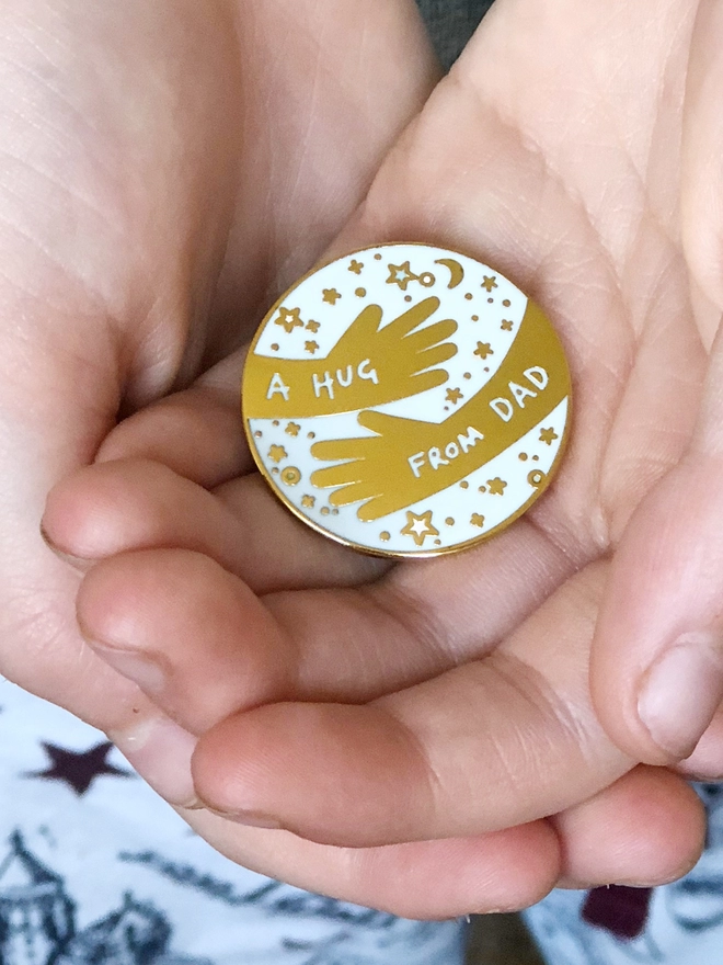 A young child holding a white and gold a hug from dad pin badge in their hands