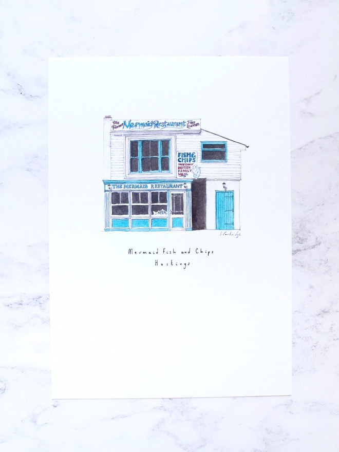 Watercolour illustration of Mermaid Fish and Chips a white and blue seaside fish and chip shop. The illustration is a small watercolour painting in the centre of the white page, the photo background is a pale white marble.