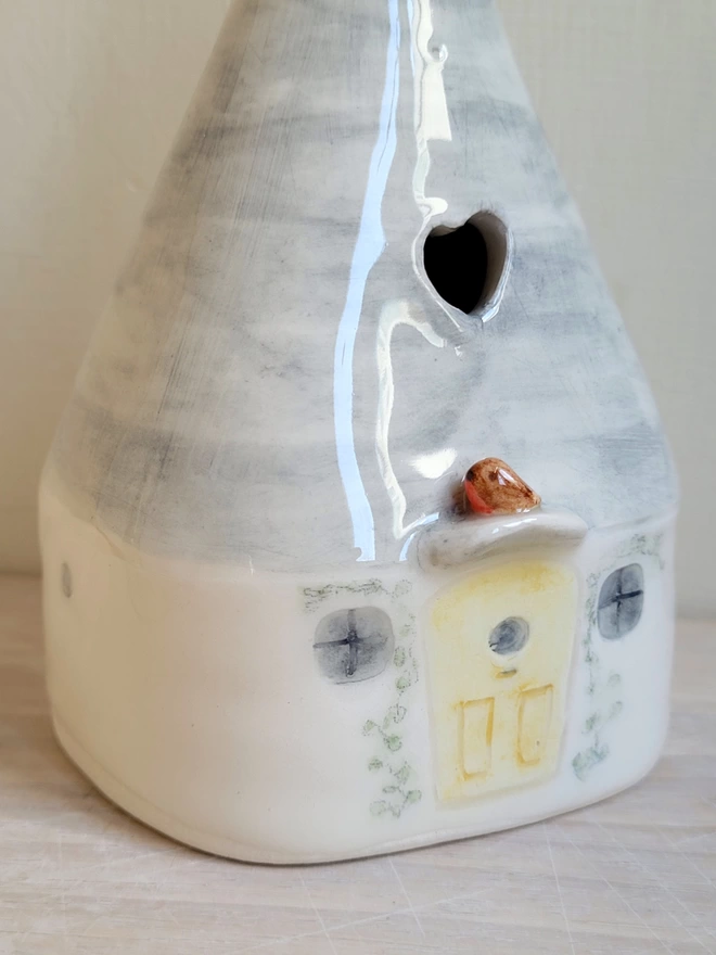 close up of a ceramic vase shaped like a house there are 2 grey windows and a yellow door with handpainted climbing plants above which is perched a modelled robin bird there is a heart cut out of the grey roof