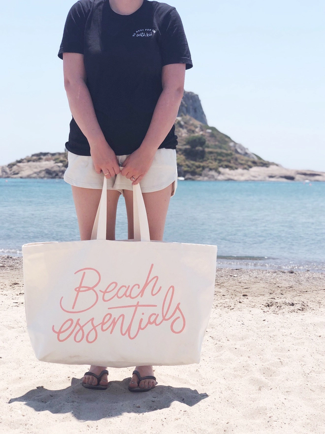 model holding a oversized natural canvas tote bag with beach essentials slogan on the beach